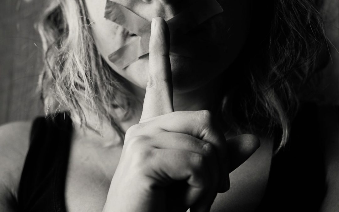 Woman holding finger to lips, mouth has tape across it