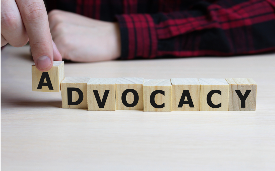 ADVOCACY spelt with wooden blocks - hand putting down letter A - arm in background wearing red checked flannel
