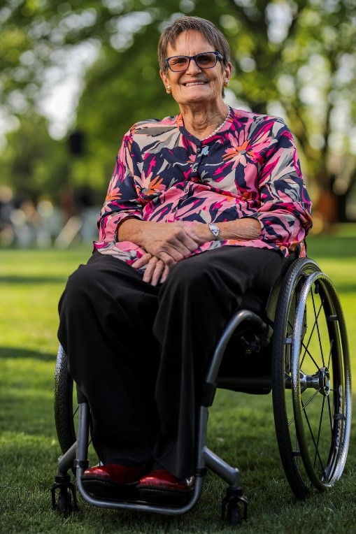 Sue Salthouse, using wheelchair, wearing bright floral top, pearl necklace and sunglasses in park